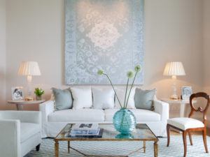 A bright living room by Susan Stacy Design features a white sofa, light blue cushions, a glass coffee table with books, large blue patterned artwork, two lamps on side tables, a white armchair, and a wooden chair.