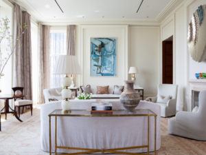 A bright, elegant living room by Susan Stacy Design features light-colored walls and furniture, large windows with sheer curtains, a sofa, armchairs, a console table with lamps and vases, and a blue abstract painting on the wall.