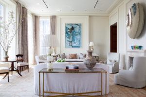 Elegant living room with neutral tones, designed by Susan Stacy Design, featuring a white sofa, armchairs, large windows with sheer curtains, modern art on the wall, decorative vases, and a gold-accented console table. Light fills the spacious area.