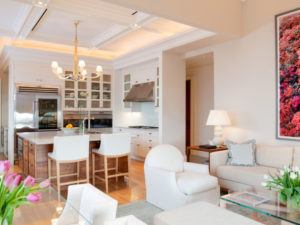 A modern, open-concept kitchen and living room by Susan Stacy Design features white cabinets, a marble island, wooden accents, cozy beige seating, tulips on the table, and a large colorful framed photo on the wall.
