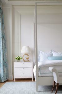 A bright, elegant bedroom by Susan Stacy Design features a white nightstand with a gold-accented lamp, a vase of green flowers, and a neatly made bed with white and light blue bedding. Blue floral curtains and a patterned rug complete the decor.