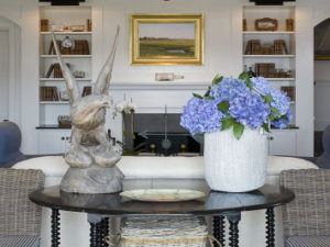 A bright Susan Stacy Design living room features a table with a wooden bird sculpture and a white vase of blue hydrangeas, set before a sofa and built-in shelves. A landscape painting hangs above the fireplace, adding a serene touch.