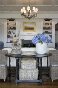A stylish living room by Susan Stacy Design features a dark wood console table with wicker baskets underneath, a large vase of blue hydrangeas, and a wooden sculpture. Built-in shelves, framed art, and a chandelier complete the decor.