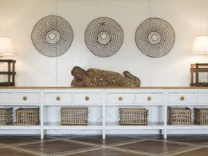 A white console table with baskets underneath by Susan Stacy Design holds a reclining statue. Two lamps sit at each end, while three decorative round wall hangings are displayed on the white wall above.