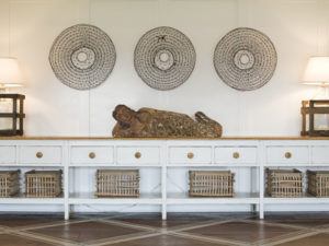 A serene, white console table with wicker baskets from Susan Stacy Design is topped by a reclining Buddha statue. Three round woven wall art pieces and two lamps with rectangular shades decorate the wall behind.
