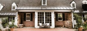 A brick house with white trim and black shutters features a central entrance with large glass doors, a bench with cushions, stacked firewood, potted plants, and easy access to Edson Hill Contact Information.
