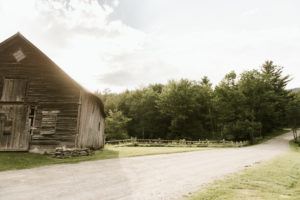 An old wooden barn sits beside a gravel road, surrounded by green grass and dense trees under a partly cloudy sky—sunlight streaming through, capturing the peaceful charm of a Vermont vacation.