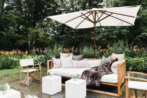A cozy outdoor seating area with a wooden sofa covered in cushions and a blanket under a large white umbrella, surrounded by greenery and flowers—perfect for relaxing during your Vermont vacation.