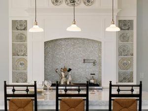 A modern kitchen by Susan Stacy Design features a marble island, three high-backed chairs, pendant lights, a tiled backsplash, built-in shelves displaying plates, and various utensils and glasses on the countertop.
