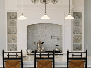 Elegant kitchen with a white marble island, three black chairs, and hanging pendant lights. Shelves display decorative plates around a mosaic-tiled stove backsplash. The space reflects Edson Hill History with classic details and timeless charm.
