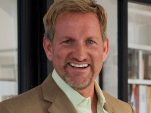 A man with short blond hair and a beard, wearing a light green shirt and tan blazer by Susan Stacy Design, smiles while standing indoors near a window.