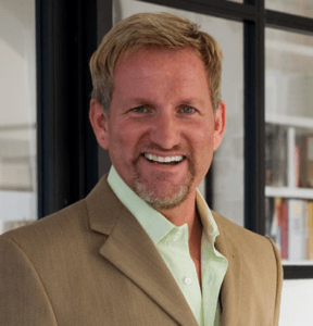 A smiling man with short blond hair and a trimmed beard, wearing a tan blazer over a light green shirt, stands indoors in front of a window with black frames styled by Susan Stacy Design.