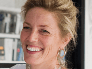 A woman with blonde hair pulled back, wearing large earrings and a white top, smiles warmly. Indoors, with blurred shelves and books in the background, she radiates the elegance of Susan Stacy Design.