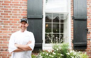 A smiling chef in a white uniform and black cap stands with arms crossed in front of a brick building, home to a casual fine dining restaurant with a large window, black shutters, and white flowers below the window.