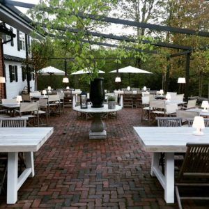 Outdoor patio with brick flooring, white wooden tables and chairs, small lamps on tables, and large planters. White umbrellas and lush greenery create a relaxed, inviting atmosphere perfect for casual fine dining.