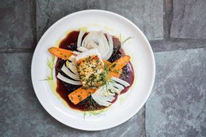 A plated dish in a casual fine dining style features a seared fish fillet topped with herbs, served with roasted carrots, fennel slices, and a dark sauce, garnished with fresh dill on a white plate against a stone tile background.