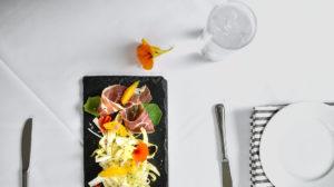 A slate platter with prosciutto, shaved fennel, orange slices, and edible flowers sets the scene for casual fine dining on a white tablecloth beside a glass of water, striped napkin, cutlery, and a white plate.