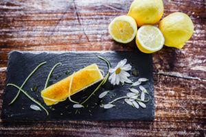 A gourmet lemon dessert on a black slate, garnished with daisy petals, green zest, and syrup—perfect for casual fine dining. Whole and halved lemons rest nearby on a rustic wooden table, enhancing the elegant yet relaxed atmosphere.