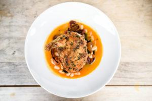 A white plate with a seasoned, cooked pork chop rests on a bed of white beans and vegetables in an orange sauce, showcasing casual fine dining from above on a wooden table.