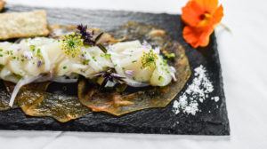 A gourmet dish perfect for casual fine dining, it features finely sliced seafood atop crispy rounds on a black slate plate, garnished with herbs and edible flowers, with salt flakes and a bright orange flower on the side.