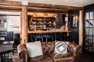 A cozy pub interior blends casual fine dining with comfort, featuring a brown leather sofa with patterned cushions, a wooden bar lined with bottles on shelves behind it, black bar stools, and rustic wood beams and paneling.