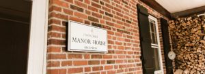 A white sign reading Edison Hill Manor House Reception is mounted on a red brick wall near a window, with stacked firewood and a black shuttered window nearby—capturing the welcoming charm found in many Stowe VT Jobs settings.