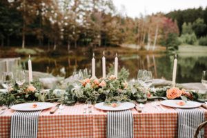 A beautifully set outdoor table with an orange checkered tablecloth, striped napkins, floral centerpieces, candles, wine glasses, and plates overlooks a calm lake with trees—a scene reminiscent of Stowe Vermont lodging charm.