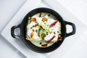 A black cast iron dish with creamy sauce topped enchiladas, garnished with pumpkin seeds, strips of dried chili, and green herbs, embodies the essence of Casual Fine Dining on a white plate and tablecloth.