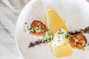 A white plate with two seared scallops, microgreens, a dollop of creamy sauce, a smear of yellow sauce, and a sprinkle of dark crumbs offers an elegant touch of casual fine dining on a light wooden table.
