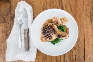 A grilled pork chop with grill marks sits atop greens and a slice of bread, topped with brown sauce, on a white plate—an inviting Casual Fine Dining presentation. A knife and fork rest on a white napkin to the left, all on a wooden table.
