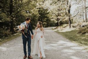 A bride in a flowing white dress and a groom in a navy suit walk hand in hand down a sunlit, tree-lined dirt road at Edson Hill - Stowe Vermont Inn. The groom holds a bouquet as they smile warmly at each other.