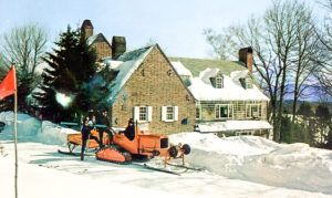A vintage snowcat vehicle plows snow in front of a large brick house with a snowy roof and chimneys, surrounded by tall trees—a perfect scene for cross country skiing on a bright winter day.