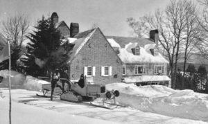 A vintage black-and-white photo shows a snow-covered house with a group of people riding a small tracked snow vehicle on a plowed path, while others prepare for cross country skiing among leafless trees under a clear sky.