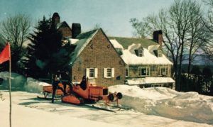 A vintage snowplow clears a snow-covered driveway in front of a large brick house with snowy rooftops and chimneys, while leafless trees hint at perfect conditions for cross country skiing on this sunny winter day.