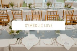 Elegant weddings reception table with wooden chairs, white linens, blue table runner, floral centerpieces, and place settings. Text overlay reads SYMBOLIC LOVE in gold letters.