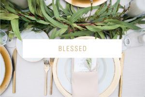 A table place setting perfect for weddings, featuring gold utensils, white plates with gold rims, a beige napkin, a green leafy centerpiece, and the word BLESSED written in gold letters across the center.