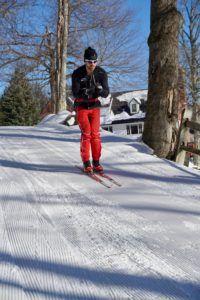 A person in red pants and a black jacket enjoys cross country skiing on a snowy, groomed trail surrounded by trees, with a house visible in the background under a clear blue sky.