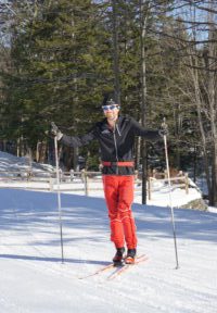 A person in red pants and a black jacket is cross country skiing on a snowy trail surrounded by trees, smiling and holding ski poles with arms outstretched.
