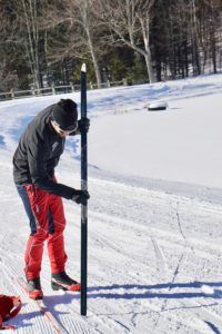 A person in red ski pants and a black jacket stands on snowy ground, holding a cross-country ski vertically, ready for a day of cross country skiing, with a forest in the background on a sunny winter day.