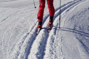 Person in red ski pants and black ski boots enjoying cross country skiing on a groomed, snowy trail, with ski poles in hand and clear parallel ski tracks visible in the snow.