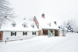 A cozy house covered in snow, with packages stacked on the decorated porch and snow-laden trees around. The scene is bright and wintry, suggesting a peaceful, festive winter day.