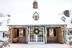A cozy brick house covered in snow, decorated with garlands and a large wreath on the glass-paned front doors. Stacks of firewood and neatly wrapped packages sit under the porch roof, while warm light shines from the windows.
