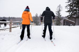 Two people dressed in winter clothing cross-country skiing side by side on a snowy trail, each carrying small packages, surrounded by trees and wooden fences on an overcast day.