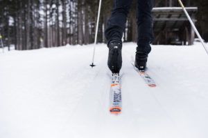Close-up view of a person cross-country skiing on a snowy trail through a forest, focusing on their skis, boots, and gear packages as they glide forward.