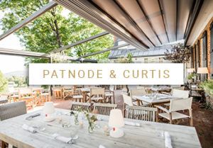 Outdoor patio dining area with wooden tables and chairs set for a meal under a partially open pergola; perfect for weddings, with green trees and blue sky in the background. Text overlay reads PATNODE & CURTIS.