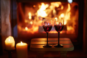 Two glasses of red wine on a small wooden table in front of a warm fireplace; two lit candles nearby create a cozy and romantic atmosphere, perfect for those seeking unforgettable romantic getaways in Vermont.