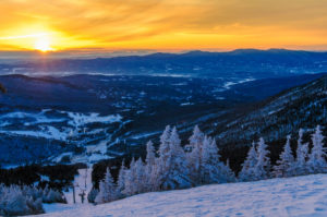 Snow-covered trees and ski slopes on a mountain overlook a valley at sunset in Stowe skiing country, with the sun casting a golden glow over distant hills and a sky filled with warm orange and yellow hues.