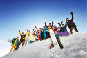 A group of snowboarders in colorful winter gear pose and wave on a snowy slope under a clear blue sky, smiling energetically as they enjoy their day after checking into cozy Stowe lodging.