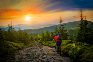 A cyclist wearing a helmet and backpack rides a mountain bike on a rocky forest trail at sunset—one of the top things to do in Stowe VT—surrounded by lush green trees and hills under a colorful sky.