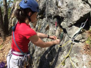 A person wearing a helmet and red shirt prepares climbing gear, adjusting ropes and carabiners attached to anchors on a rocky outdoor climbing route, ensuring all safety packages are secure before the ascent.